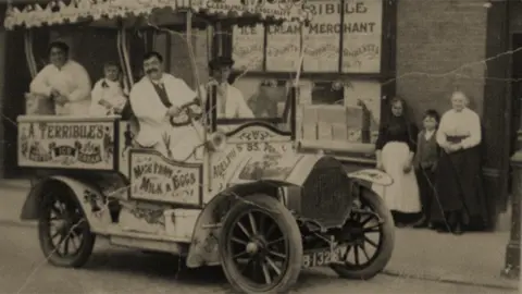 Edwin Williams Terribile family with their motorised ice cream vehicle in 1917. There are four people smiling in the open top car all wearing white jackets and they are parked outside the shop watched by two women in long skirts and aprons with a small boy between them