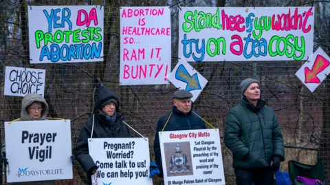 PA Media Pro-life protesters stand in zipped up coats and hats and hoods holding signs urging women to rethink abortion choices. Neon hand-made pro-choice signs in counter protest are hung on a fence behind them.
