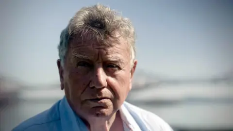 Rogan Scotland George Thomson is looking into the camera with a serious face. He has short, grey hair and is wearing a white shirt. 