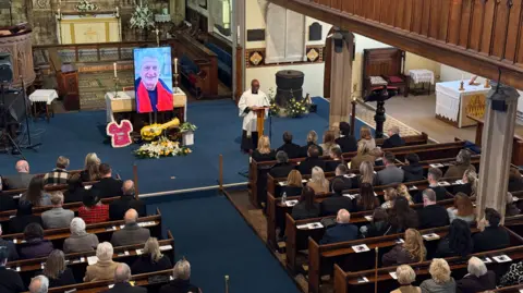 A large picture of a man, with tributes underneath it, is placed at the front of a church. People are in a rows of pews, looking towards the front.