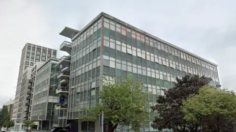 A grey, glass city-centre office block. There are trees in front of the large building. 