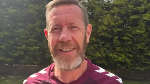 Colin Brown Colin Brown has short fair hair and smiles at the camera. He wears a maroon Hearts FC strip and is standing in a garden with a bush behind him
