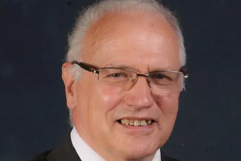 Fife Council A man with receding grey hair and glasses in a dark suit jacket with a white shirt and red tie
