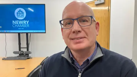 A man in a blue fleece with a blue shirt. He wears glasses. There is a desk behind him and a TV.
