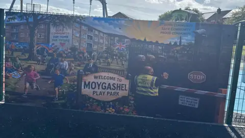 Banner on a play park fence, showing children playing sunshine and men being rejected at a barrier. 