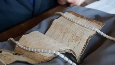 Isabelle Freeman An old manuscript is lying open on a table. The pages and sleeve are very old and worn and the writing is not legible
