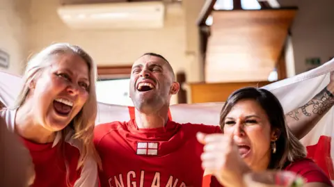 Two women and a man celebrate. They are in a pub wearing England football shirts and holding a flag
