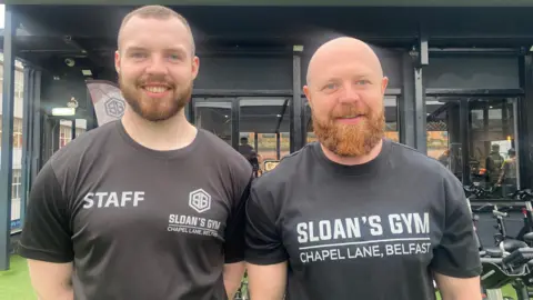 Curtis and Johnny Sloan smile at the camera, standing on the gym rooftop. They are wearing black T-shirts. Both have brown beards. Behind them is the gym. Peleton bikes are outside. There is artificial grass. The gym building is painted black. 