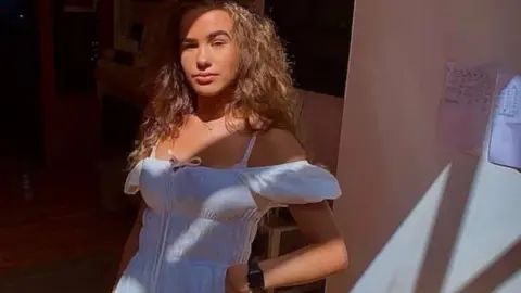 Brooke George is looking at the camera wearing a cotton off-the-shoulder dress. She has long wavy blonde hair and is posing for the camera with her hand on her hip