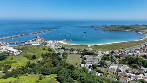 Aerial view of a coastal town with a harbour full of small boats, a curved sandy beach, and houses surrounded by green landscape under a clear blue sky.
