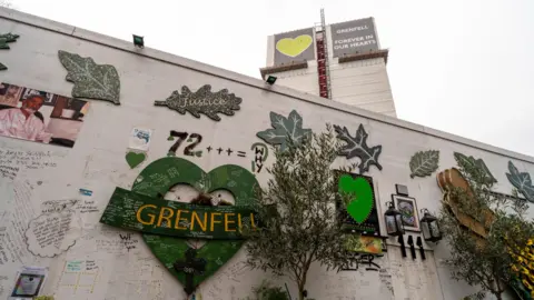 PA Media A file image showing Grenfell Tower covered in tarpaulin and a grey and green sign with a heart saying 'Grenfell: Forever in our hearts' in the background while white hoarding sits in the foreground. A green heart with the word 'Grenfell', green leaves, picture and handwritten tributes cover the hoarding