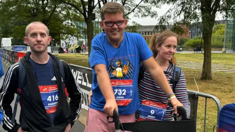 John Noble crosses the finish line with a walking frame. He has two friends flanking him, a woman in a stripey top and a man wearing a rucksack. Each has a red and blue label with a race number on them, they read 416 Andrew, 732 John, and 9 Jenny.