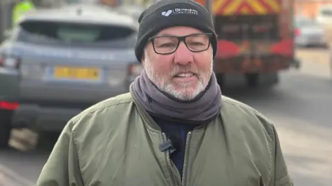 BBC A middle-aged man wearing a black woolly hat with Birmingham City Council written on the front smiles into the camera. He is wearing black-rimmed glasses, a scarf and a green jacket.