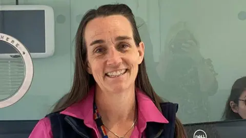 Katie Lennon is smiling at the camera. She has long brown hair and is wearing a pink shirt.