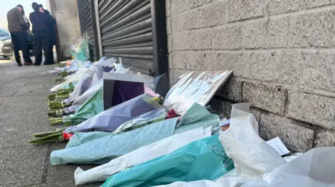 About 20 bouquets of flowers, some with messages attached, in a row on the ground outside a grey, brick-built building with black shutters. In the background, people can be seen standing on the pavement.