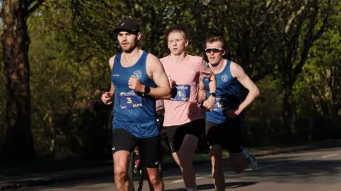 Three men running the Hospice Half-Marathon. Two are wearing blue vests and black shorts, and the third is wearing a pink top and black shorts.