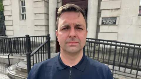 Conradh na Gaeilge Conchúr Ó Muadaigh, an man with short, brown hair, stands outside a court building, facing the camera, with a serious expression. He is wearing a navy polo shirt with a zipped neck fastening. 