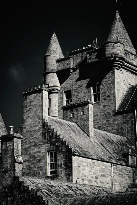 Graeme Baxter Black-and-white image of a stone building with pointed turrets, sloping roofs, and tall windows. Moonlight highlights parts of texture on the stone walls and rooftops, while deep shadows and a dark sky create a dramatic atmosphere. 
