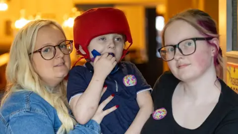A blonde woman in a denim jacket and a teenage girl with glasses stand on either side of a young boy, both holding him up between them. The boy is wearing a red protective helmet and a navy top, and is holding something to his mouth. They are inside what appears to be a restaurant or function room. 