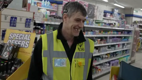 Dave Dixon is inside Boyes as sheleves of goods are filled behind him. Dave is smiling. He is wearing a yellow high vis unifom with a name card stuck to the right of his jacket.