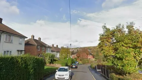 A screenshot from Google streetview of Barlow Drive, Sheffield. There is a green, leafy tree to the right and on the left is a row of semi-detached homes with front gardens. Two cars are also parked on the road.