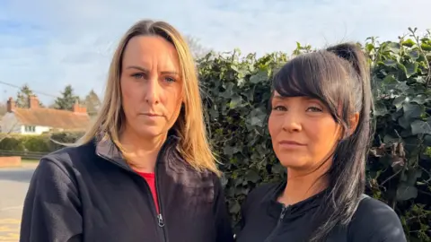 Two women looking straight at the camera. They are both wearing black zipped up jackets and standing in front of a hedge.