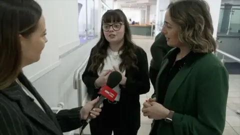 BBC journalist interviews two women. They are all wearing suits.