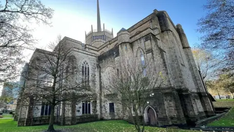 Blackburn Cathedral is a large imposing stone building set within a grassed area and surrounded by trees.