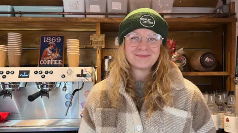A woman with blond curly hair wearing a green beanie hat saying Howley Coffee stands in front of a coffee machine