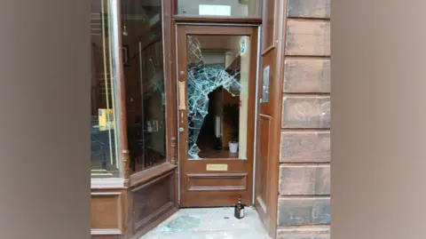 A brown framed door with glass in the centre that has been smashed. Broken glass lies on the concrete step outside the door with a bottle next to it. 