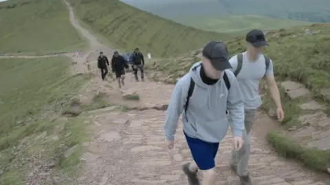 Telegram Six Active Club members, wearing sportswear and caps, walk up a stone pavement on the side of a hill, with a green valley stretching out below them