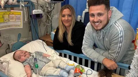 Addenbrooke's Hospital Teddy is lying in a hospital cot in Picu. He is wearing a blue and white striped top, pale joggers and blue socks. His feeding tube is taped to his left cheek, and you can see his tracheostomy tube coming out from under his chin and attached to a transparent tube with green joints. His mum and dad are leaning on the side rail of the cot and smiling. They are surrounded by machinery and a blue hospital curtain is behind them. 