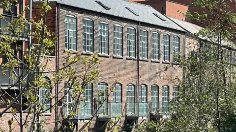 A brick built building has large glass windows with small glass balconies overlooking trees