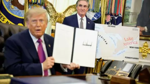 US President Donald Trump holds up a document with his signature in the Oval Office. Begin him is Secretary of the Interior Doug Burghum and a large map showing the US and Canada and the Great Lakes, with Lake Ontario renamed in red script as Lake America. 