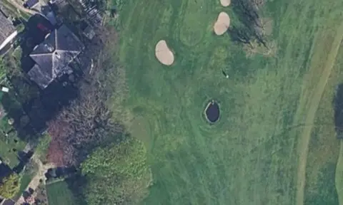 Google An aerial picture of a golf course with trees and houses bordering the site. 