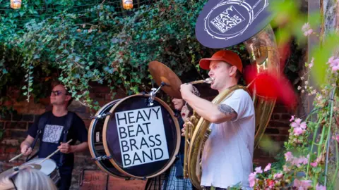 Jakeoncam Members of a band, one of whom is carrying a bass drum with a sign saying 'Heavy Beat Brass Band". A man in a red cap is playing a brass instrument and a man in the background is playing a drum. 