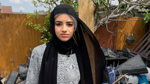 Razan, in a black headscarf, stands in her back garden surrounded by bags of compost and cannabis growing detritus