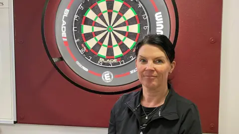 James Grant/BBC Emma Nicholls is wearing a black top and jacket, and she has black hair. She smiles in front of an illuminated darts board.