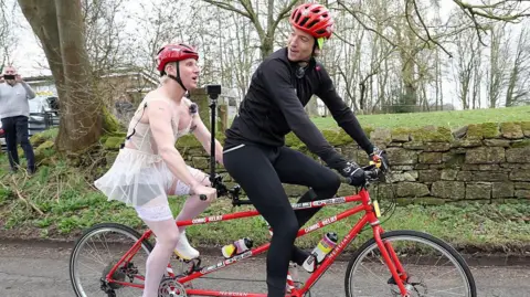 Getty Images Greg in all black cycling gear on the front seat of a red tandem bike, wearing a red helmet. Jamie is on the back seat wearing white lingerie and white stockings an a red helmet.  