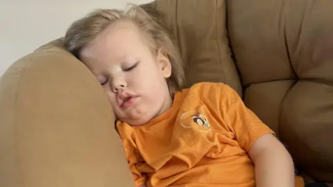 Family handout A young boy is sleeping on a beige sofa. He has blond hair and is wearing an orange t-shirt. 