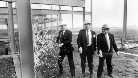 Mirrorpix/Getty Images John Hall and two other men with dark suits and ties and light shirts with hard hats on. They are next to girders on a building site. Hall is smashing a bottle of champagne on one of the girders. It is a black and white photo.