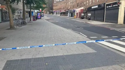 A cordoned off street with police tape sectioning off a road. There are closed shops on either side of the road in the background. There are also some trees and bicycle racks on the left side pavement in the distance. 