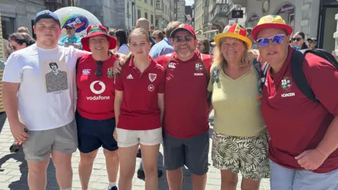 Welsh fans Aneurin Williams, Julie Cashell, Anwen Williams, Stuart Williams, Margaret Rees and Linda Rosser stand in a line with their arms around each other, smiling at the camera. Four of them are wearing red football shirts and Wales bucket hats. They are standing in a street with crowds of people behind them. 