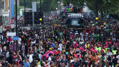 Reuters Thousands of people take part in a carnival procession along a crowded city street, with a music truck travelling through the centre of the route beneath trees.