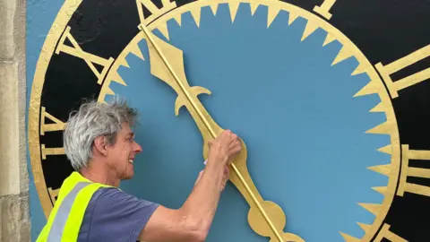 Image shows man with grey hair wearing a purple T-shirt and a bright yellow jacket applying gold leaf to the middle of the clock hand which is on bright blue background and yellow Roman numerals around the clock face