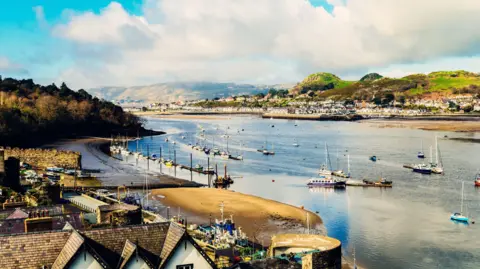 Getty Images Looking north east from the town of Conwy across the bay. There are a number of sailing boats on the water with hills and houses in the background.