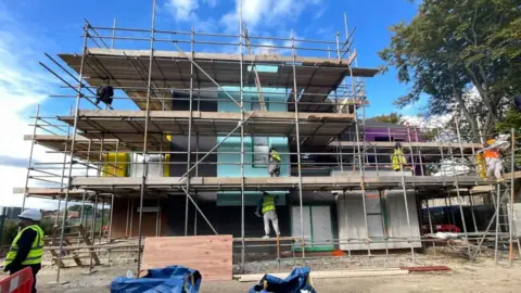 An image of a brightly coloured community centre with scaffolding around the exterior. Five workers, wearing hard hats and yellow clothing, are helping with the construction of the building.