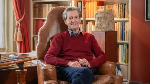 Amy Bateman A man with grey hair sitting in a brown leather chair with his hands in his lap. He is wearing a red jumper and sat in front of a bookshelf and stone bust. 