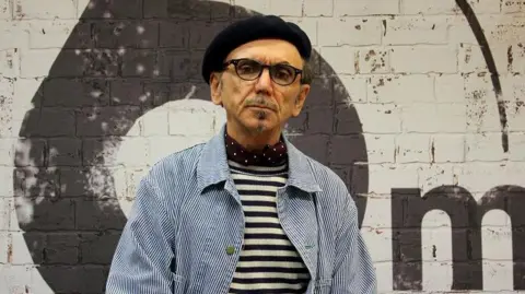 Kevin Rowland has a black goatee and short dark hair, he is wearing a black beret hat. He is also wearing a black and white striped and blue denim jacket with a burgundy scarf with white dots.
