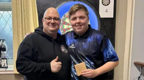 A teenager wearing a blue darts shirt stands next to a bald man in a black hoodie with his arm around his shoulder. A dart board is partially obscured on the wall behind them. Both are smiling at the camera and the man is giving a thumbs up. The boy is holding a small brown envelope.